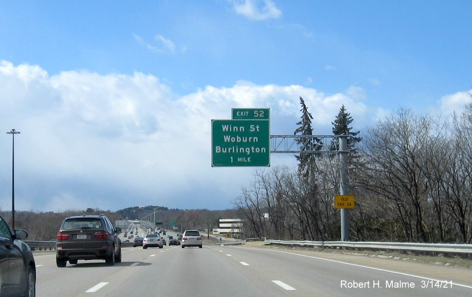 Image of 1-Mile advance overhead sign for the Winn Street exit with new milepost based exit number and yellow Old Exit 34 advisory sign on support on I-95/MA 128 South in Woburn, March 2021