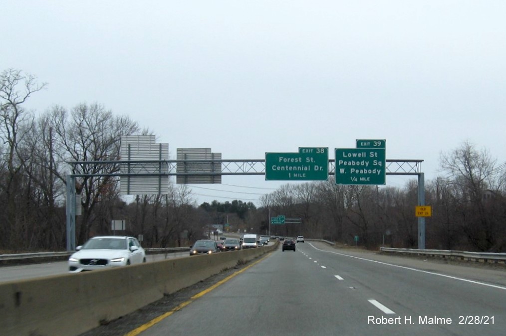 Image of overhead advance signs for Lowell Street and Forest Street/Centennial Drive exits with new milepost based exit number and yellow Old Exit 26 sign on right support post on MA 128 South in Peabody, February 2021